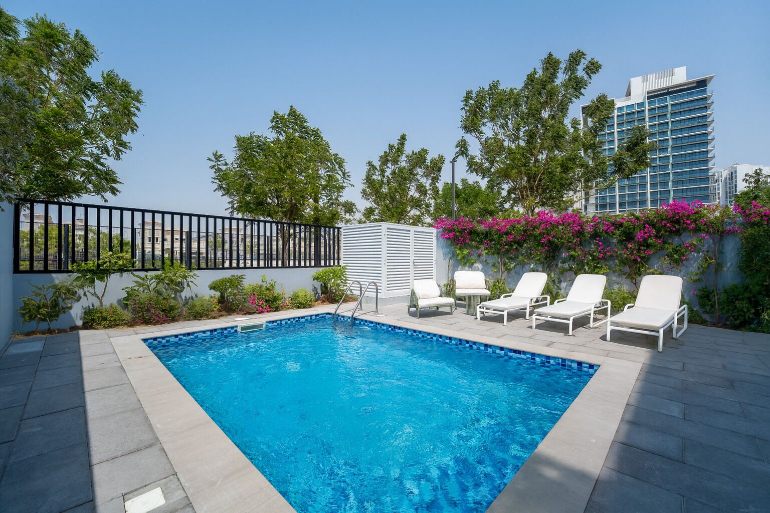 A rectangular swimming pool with a tiled edge, surrounded by a paved deck. There are four white lounge chairs and a small table with a white cushion. A metal fence encloses the area, and a building with a glass facade is visible in the background.