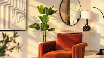 A modern living room with a plush orange chair, a large green plant, a round mirror, and a table with a lamp.