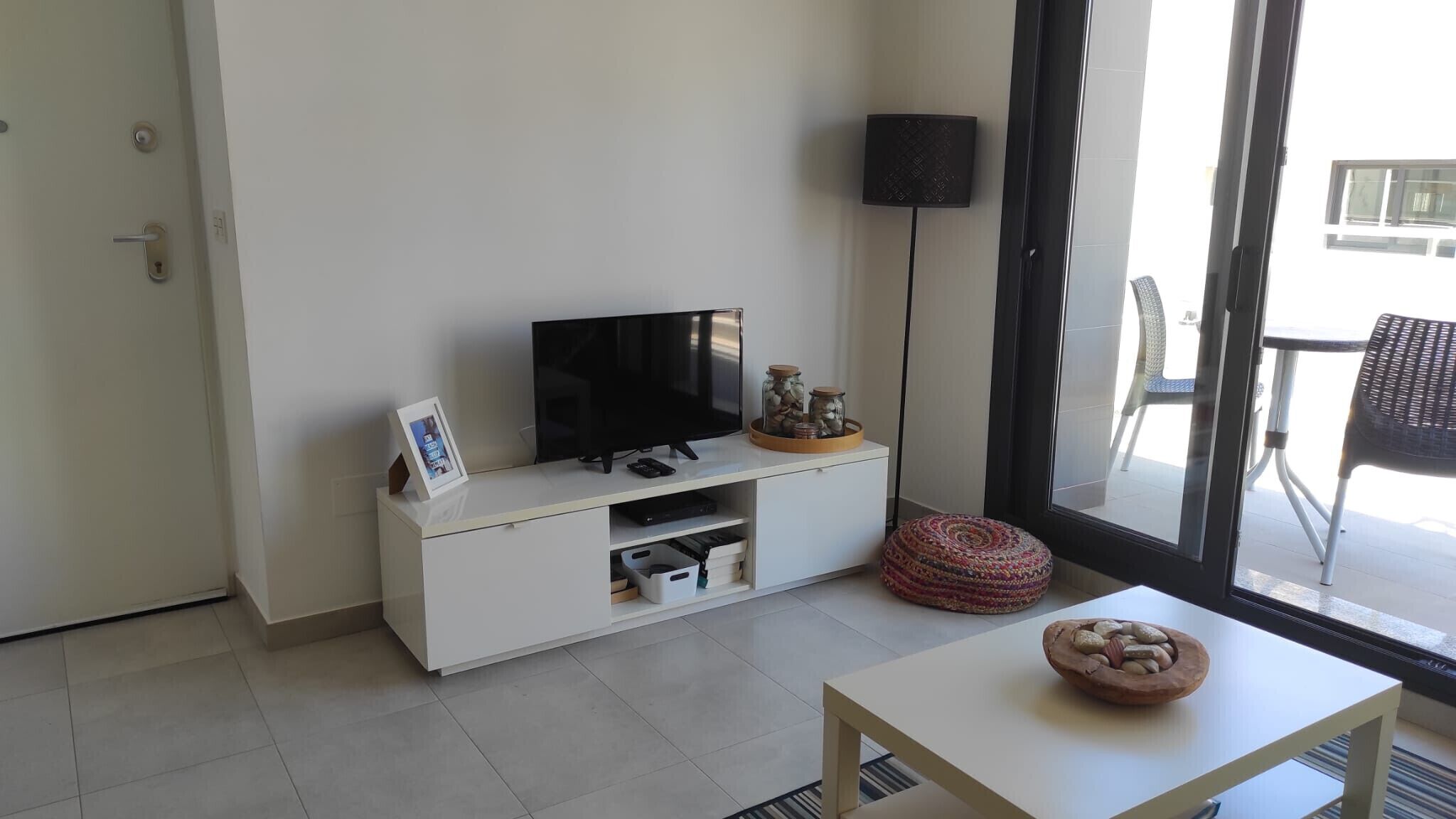 A modern living room with a flat-screen TV on a white cabinet, a small table with a bowl, and a floor lamp.
