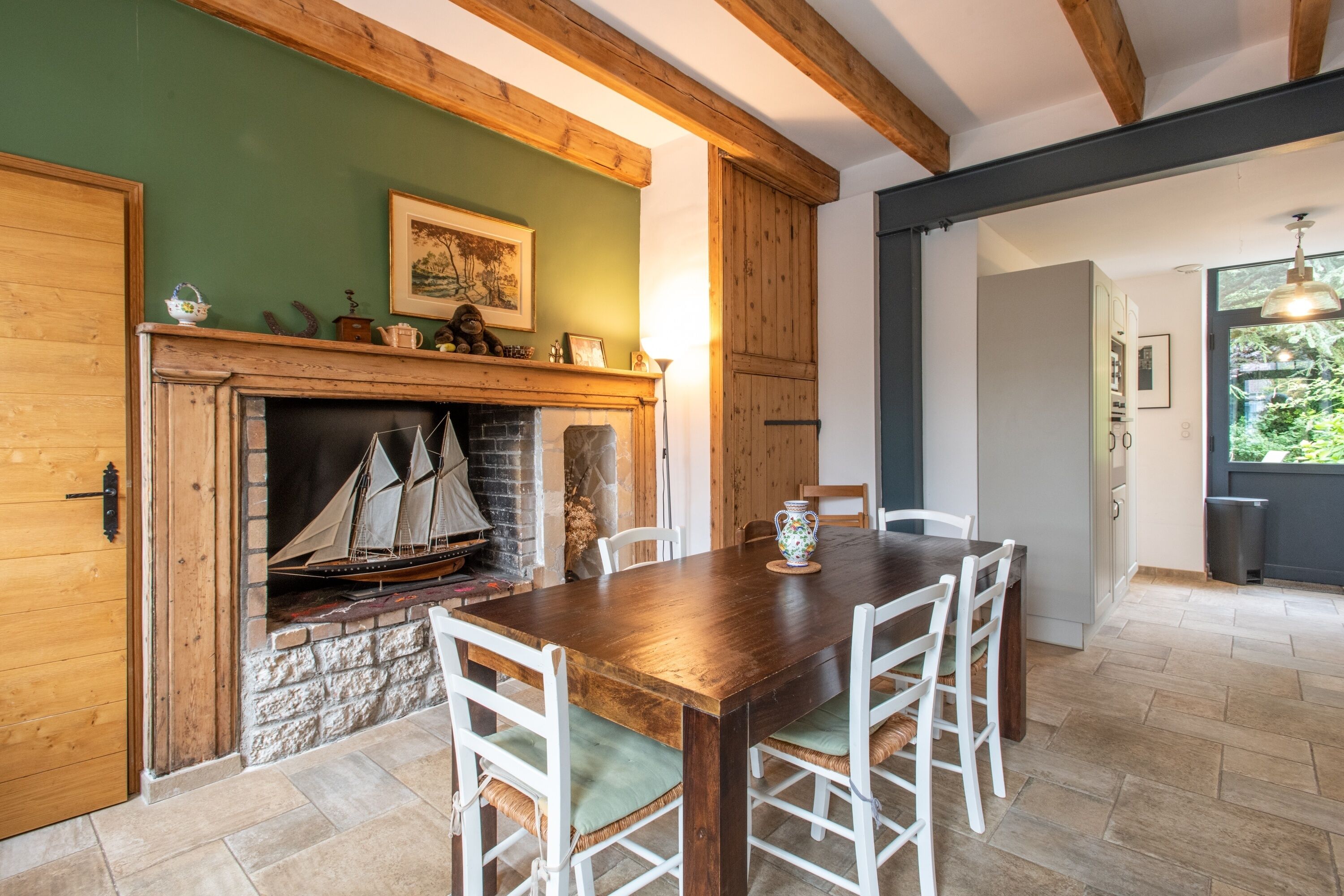 Un coin repas avec une table en bois et des chaises blanches, une cheminée ornée d’une maquette de bateau, et un mur vert agrémenté d’un tableau encadré.
