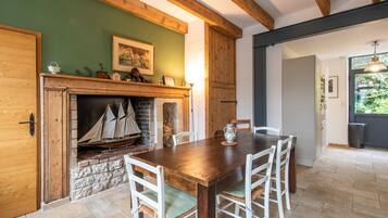 Un coin repas avec une table en bois et des chaises blanches, une cheminée ornée d’une maquette de bateau, et un mur vert agrémenté d’un tableau encadré.