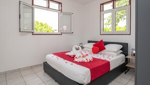 Een net opgemaakt bed met een rood-witte sprei, een handdoek in de vorm van een hart en rozenblaadjes. Twee ramen met witte kaders en open luiken, en een nachtkastje met een lamp.