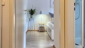 Een smalle gang met houten vloer die leidt naar een slaapkamer met een bed en nachtkastje.