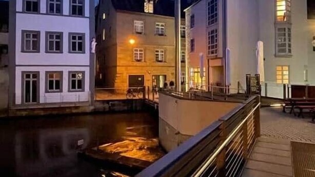 Een gebouw aan het water met een verlicht beeld in het midden, een brug met een leuning, en een rustig waterlichaam.