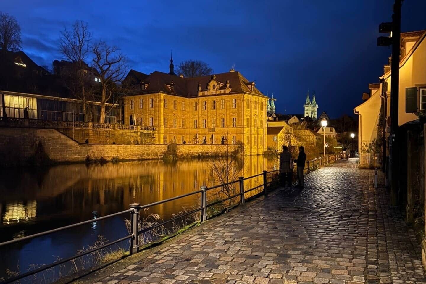 A historic building with a river in front, a cobblestone path, and a bridge.