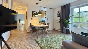 Een moderne woonkamer met een flatscreen-tv, een eethoek met een tafel en stoelen, een groot raam met gordijnen en een groen vloerkleed.