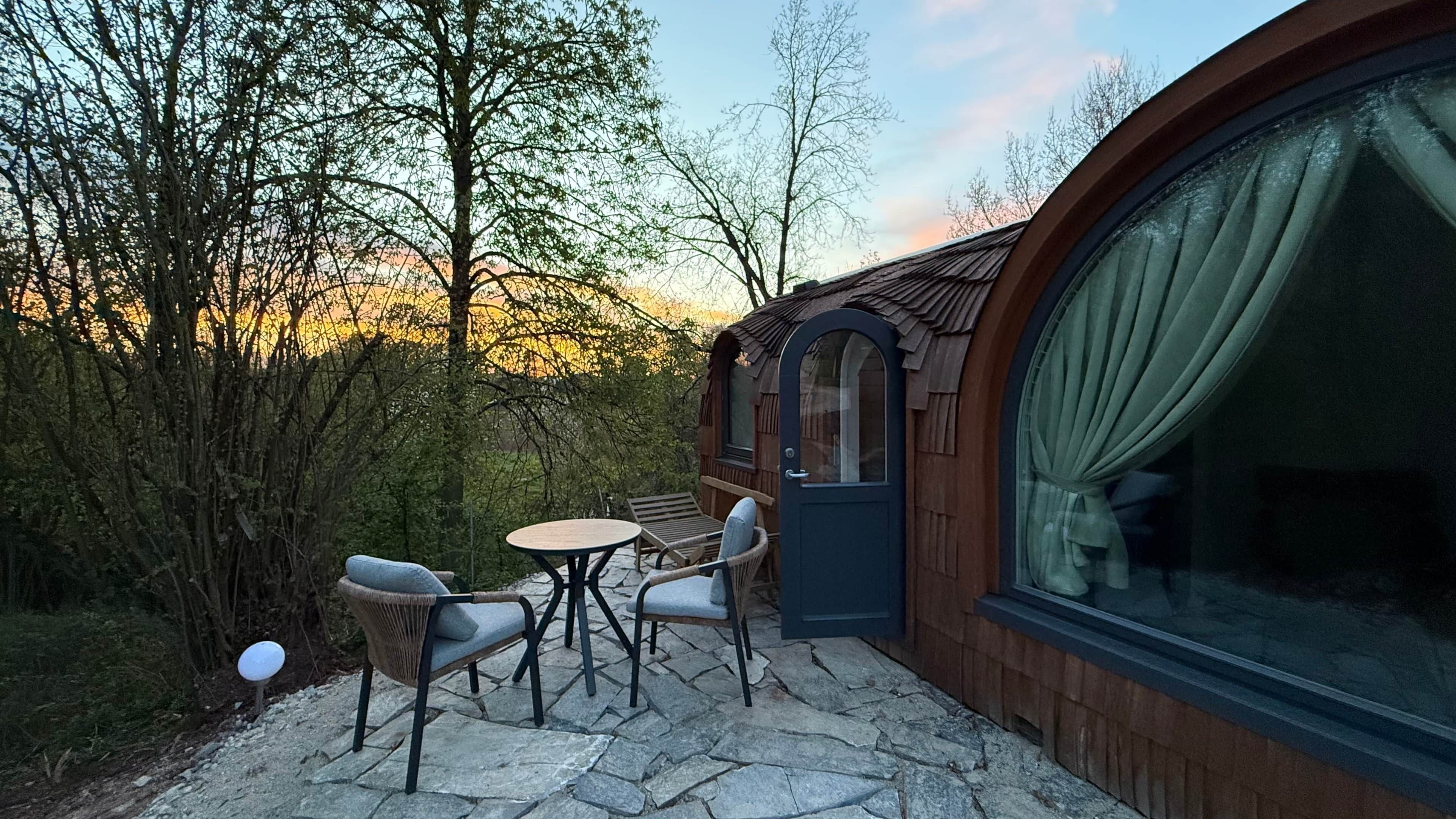 Un espace extérieur accueillant, aménagé avec une table en pierre et deux chaises, entouré d’arbres et bordé d’un bâtiment en bois doté d’une grande fenêtre.