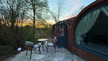 Un espace extérieur accueillant, aménagé avec une table en pierre et deux chaises, entouré d’arbres et bordé d’un bâtiment en bois doté d’une grande fenêtre.