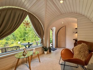 A cozy room with a curved ceiling, a wooden door, a glass table with a picture frame, a brown chair with a fluffy pillow, and a view of greenery outside. - Iglu – Chestnut (Frensdorf)