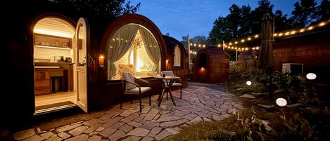 A cozy outdoor seating area with a stone-tiled patio, surrounded by wooden structures with large windows and string lights.