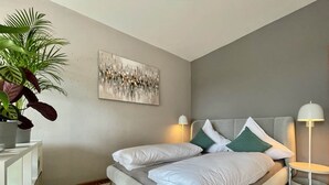 A modern bedroom with a grey bed, white pillows, a green accent pillow, a white bedside table, a floor lamp, and a large abstract painting on the wall. - Pretty City - Tiny Apartment Bamberg (Bamberg)