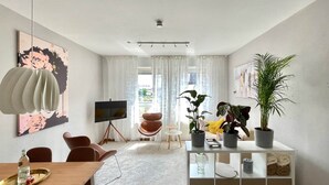 A modern living room with a TV on a tripod, a dining table with a tray, and a white sofa. There are potted plants and a white cabinet with shelves. - Pretty City - Tiny Apartment Bamberg (Bamberg)