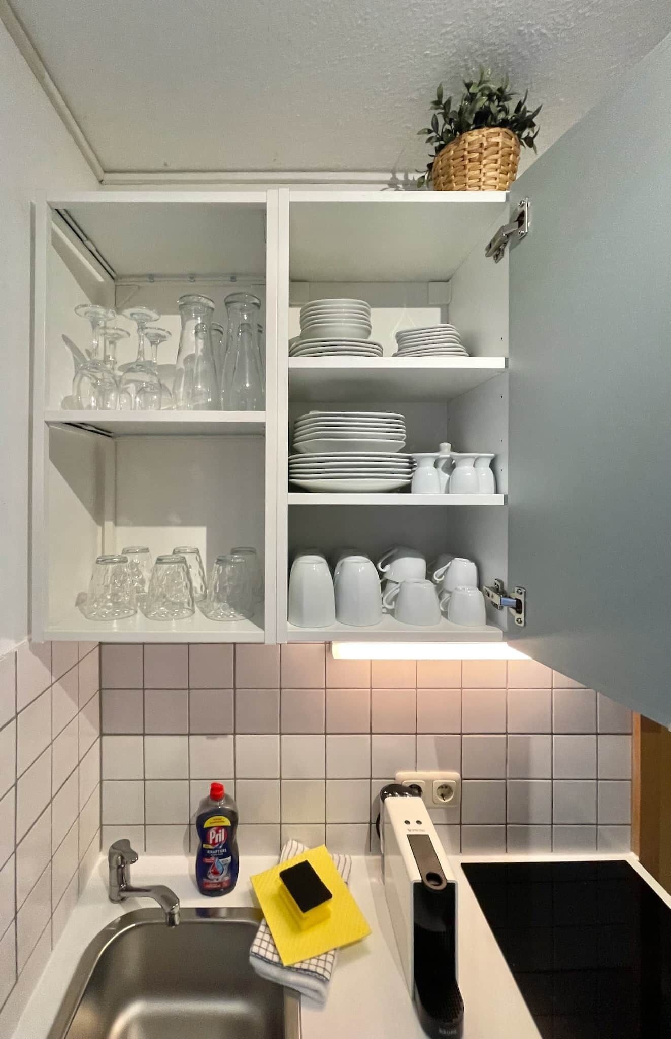 A kitchen with a white cabinet containing glassware, plates, and mugs. There is a sink with a dish soap bottle, a yellow sponge, and a black appliance.