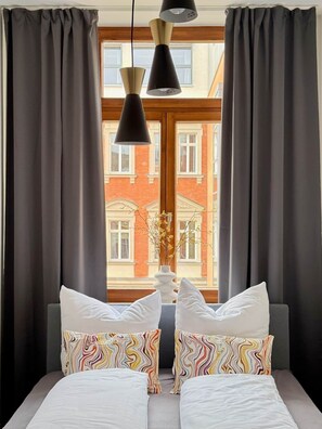 A bedroom with a large window, dark curtains, a bed with white bedding, colorful pillows, and pendant lights. - Lovely Lou - Old building apartment Bamberg (Bamberg)