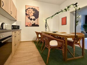 A modern kitchen with wooden cabinets, a dining area with wicker chairs, and a large painting on the wall. - Lovely Lou - Old building apartment Bamberg (Bamberg)