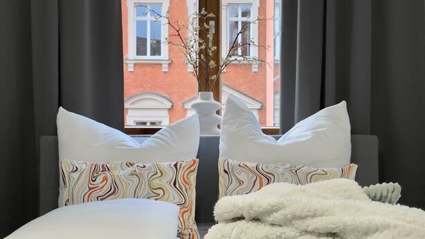 Un lit avec une literie blanche et des coussins colorés, une couverture blanche et moelleuse, et un vase avec des branches posé sur un rebord de fenêtre.