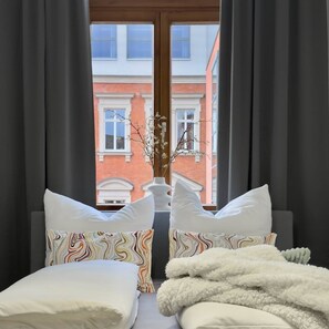 A bed with white bedding and colorful pillows, a fluffy white blanket, and a vase with branches on a windowsill. - Lovely Lou - Old building apartment Bamberg (Bamberg)