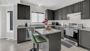 A modern kitchen with dark cabinets, a central island, stainless steel appliances, and a window with blinds. - 100% Private and Spotless ~Fresh Linens ~New House (Thorold)