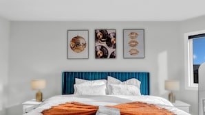 A modern hotel room with a large bed, white bedding, a blue headboard, and three framed artworks on the wall. - 100% Private and Spotless ~Fresh Linens ~New House (Thorold)