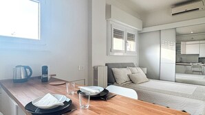 Habitación de hotel moderna y compacta con mesa de comedor de madera, cama con edredón blanco, kitchenette y ventana que proporciona luz natural.