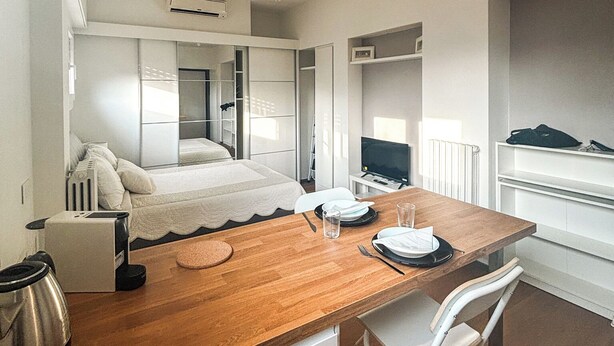 Habitación de hotel moderna y funcional con una mesa de comedor de madera, sillas blancas, una cama con edredón blanco y un televisor sobre una mesita.