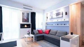 Ein modernes Wohnzimmer mit einem grauen Ecksofa, einem kleinen weißen Beistelltisch und einem weißen Couchtisch. Ein Bücherregal mit Dekorationselementen und ein Gemälde an der Wand sind ebenfalls zu sehen.