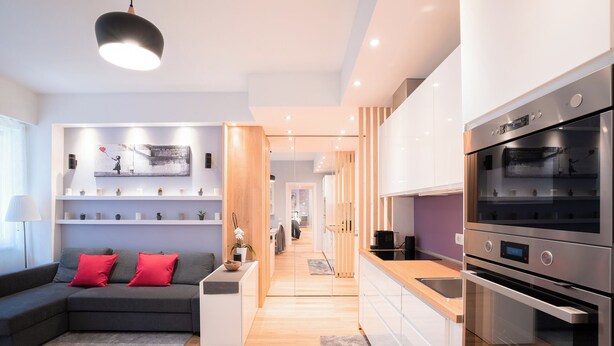Eine moderne Küche mit integrierten Geräten, ein graues Sofa mit roten Kissen und ein Essbereich aus Holz.