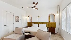 Une chambre d’hôtel comprenant un lit, deux fauteuils, une petite table, un ventilateur de plafond, un miroir et une fenêtre avec des stores.