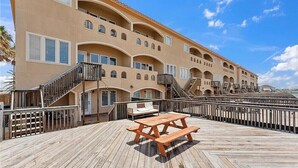 Un hôtel à plusieurs étages doté de balcons, d’une terrasse en bois avec une table de pique-nique, et un ciel d’un bleu limpide.