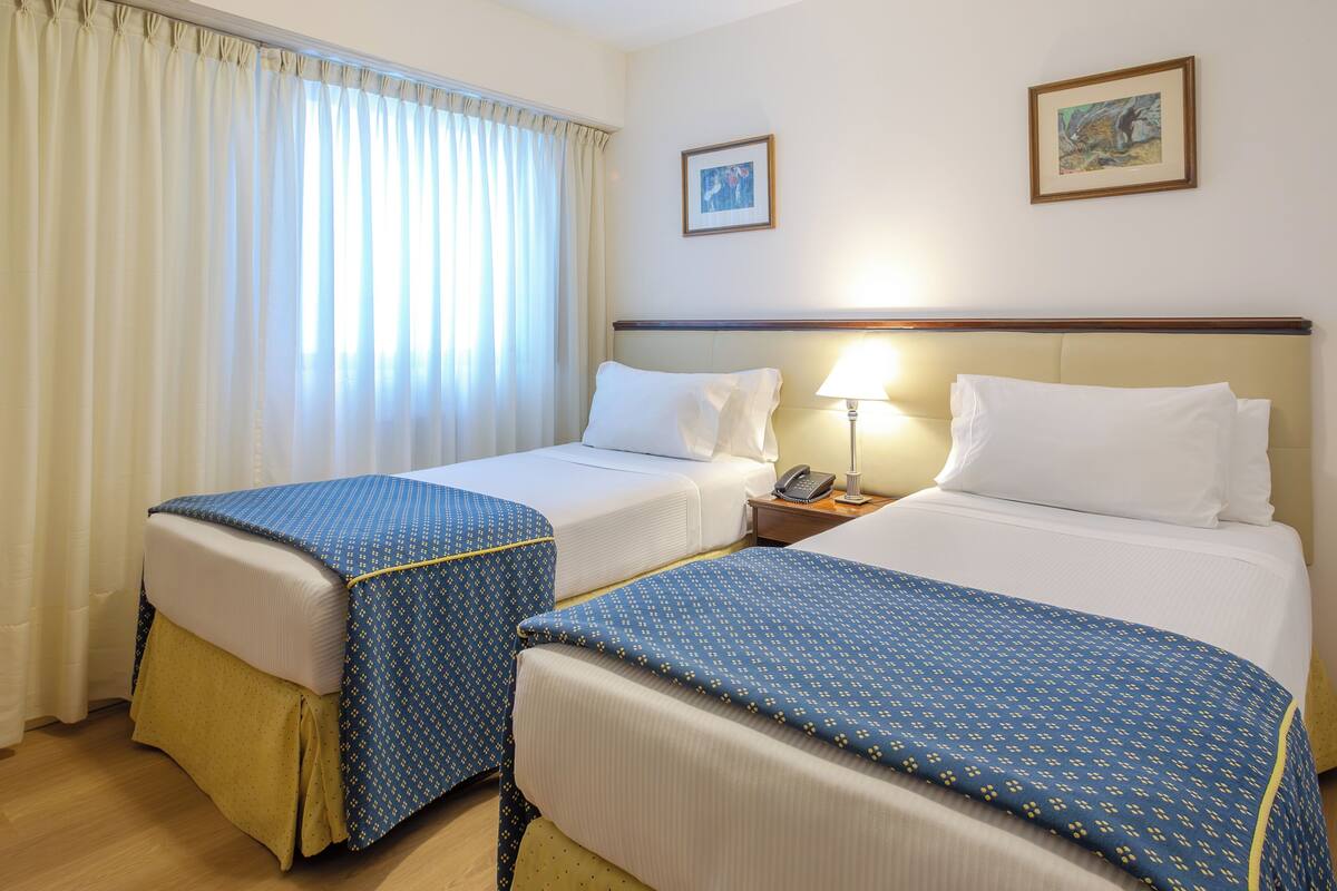 A hotel room with two beds, a nightstand with a lamp, a telephone, and a painting on the wall.