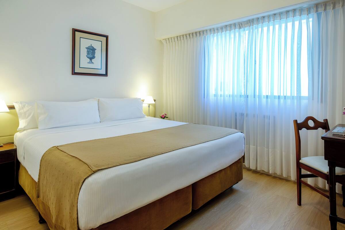 A hotel room with a large bed, a desk with a chair, a framed picture on the wall, and a window with curtains.