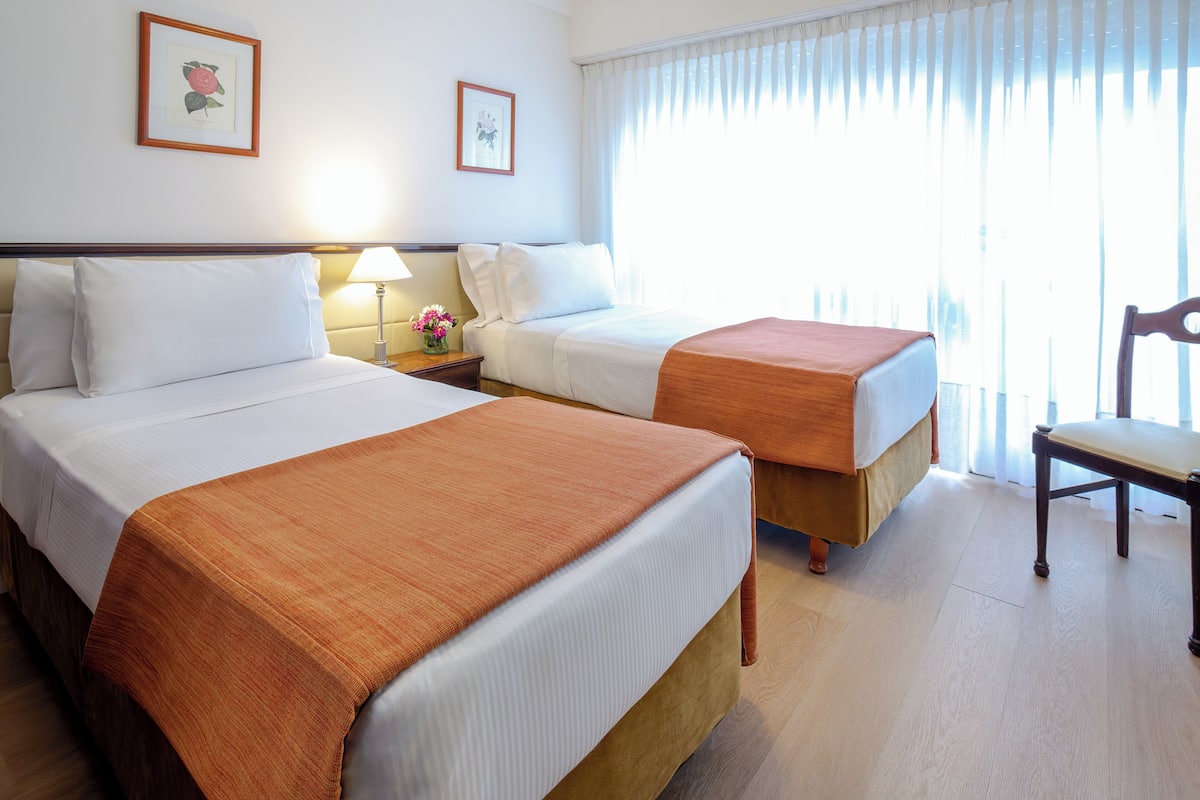 A hotel room with two beds, a nightstand with a lamp, a chair, and a window with curtains.