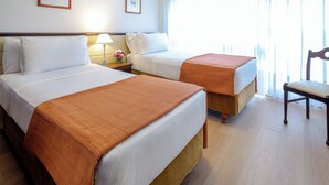 Habitación de hotel con dos camas, una mesita de noche con lámpara, una silla y un ventanal con cortinas.
