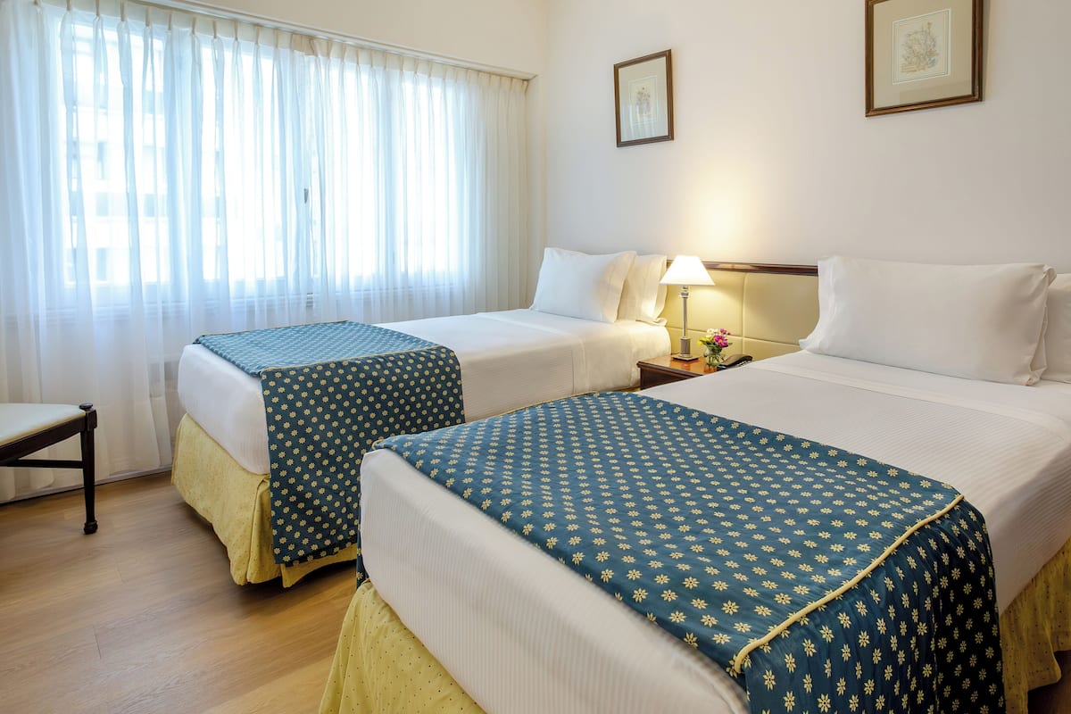 A hotel room with two beds, a chair, a nightstand with a lamp, and a window with curtains.