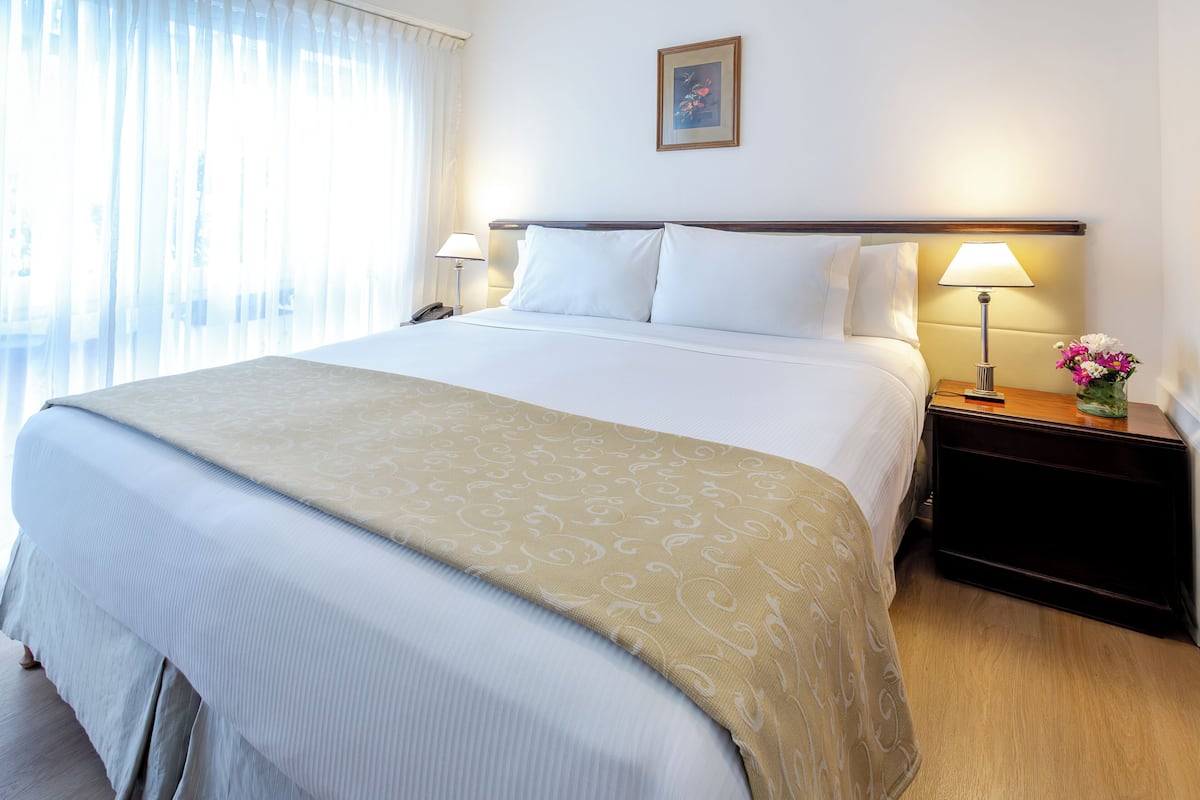 A hotel room with a large bed, two bedside tables with lamps, a nightstand with a vase of flowers, and a painting on the wall.