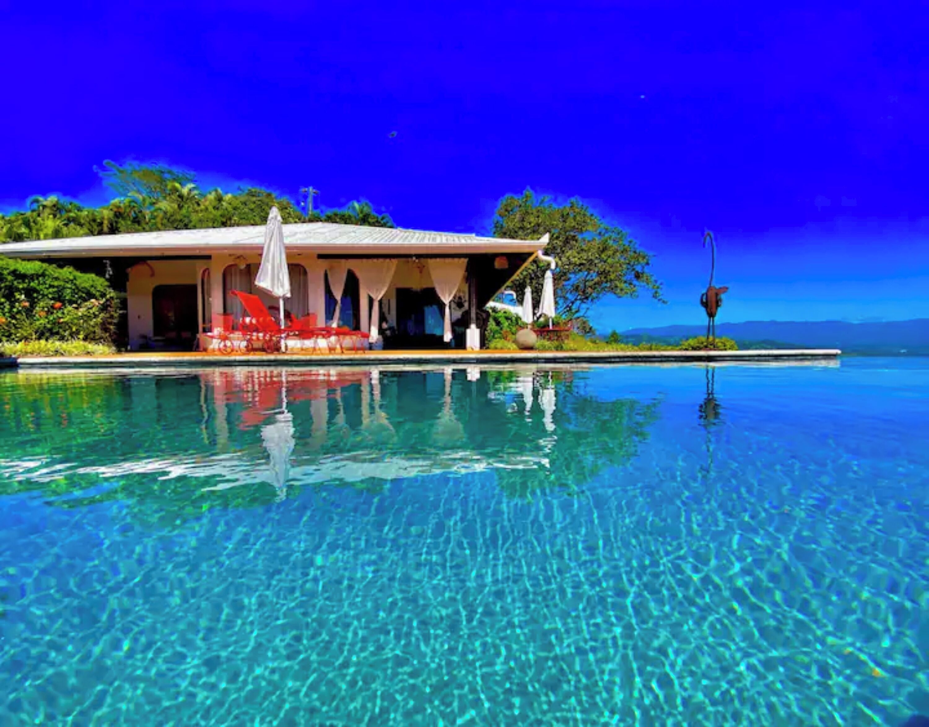 Une piscine d’un bleu éclatant, dotée d’un espace de détente et de parasols, entourée d’une végétation luxuriante et surplombée d’un ciel dégagé.