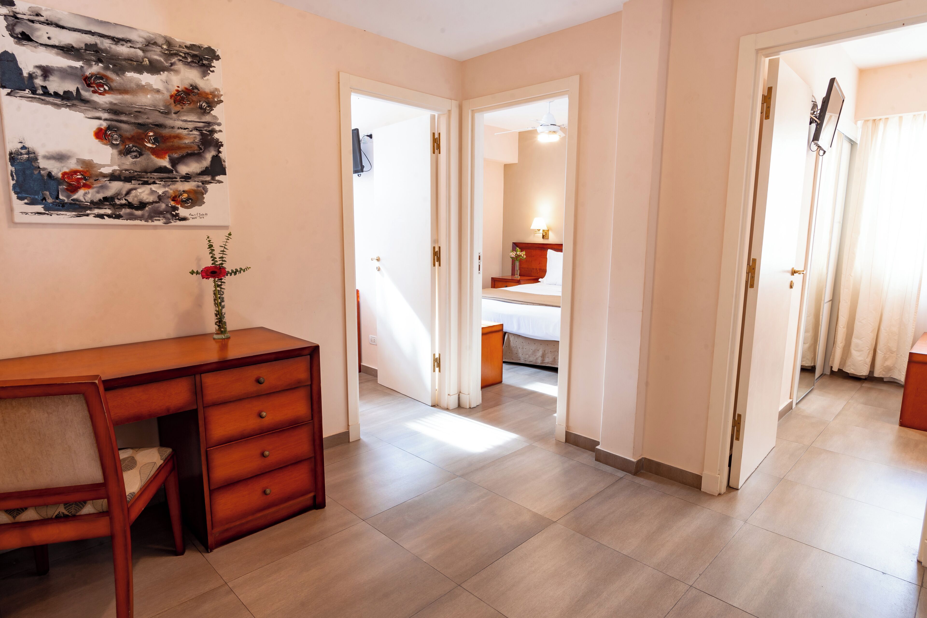 A hotel room with a wooden desk, a mirror, a bed, and a door leading to another room.