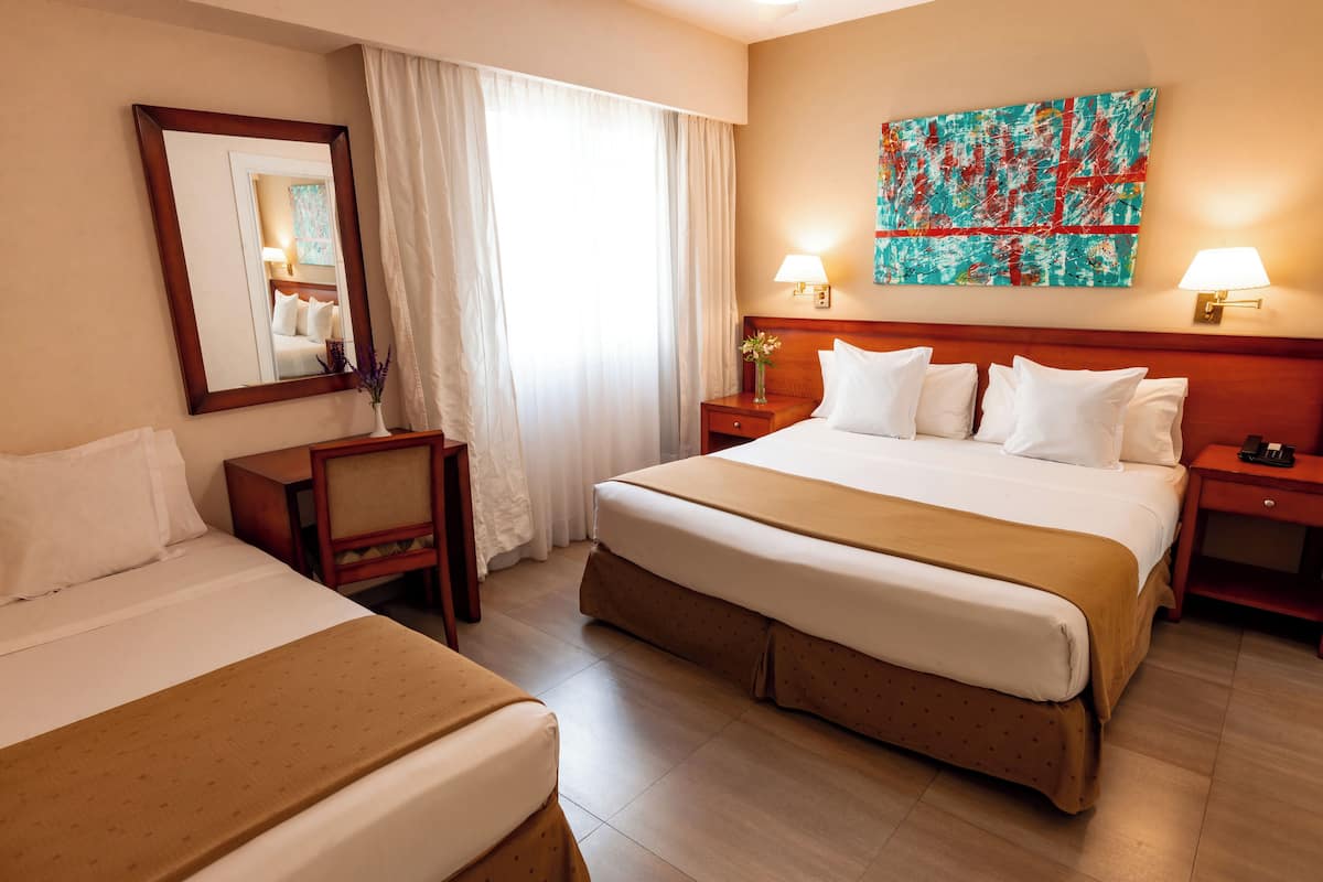 Une chambre d’hôtel avec un grand lit, un lit plus petit, une table de chevet avec une lampe, un miroir et un tableau au mur.