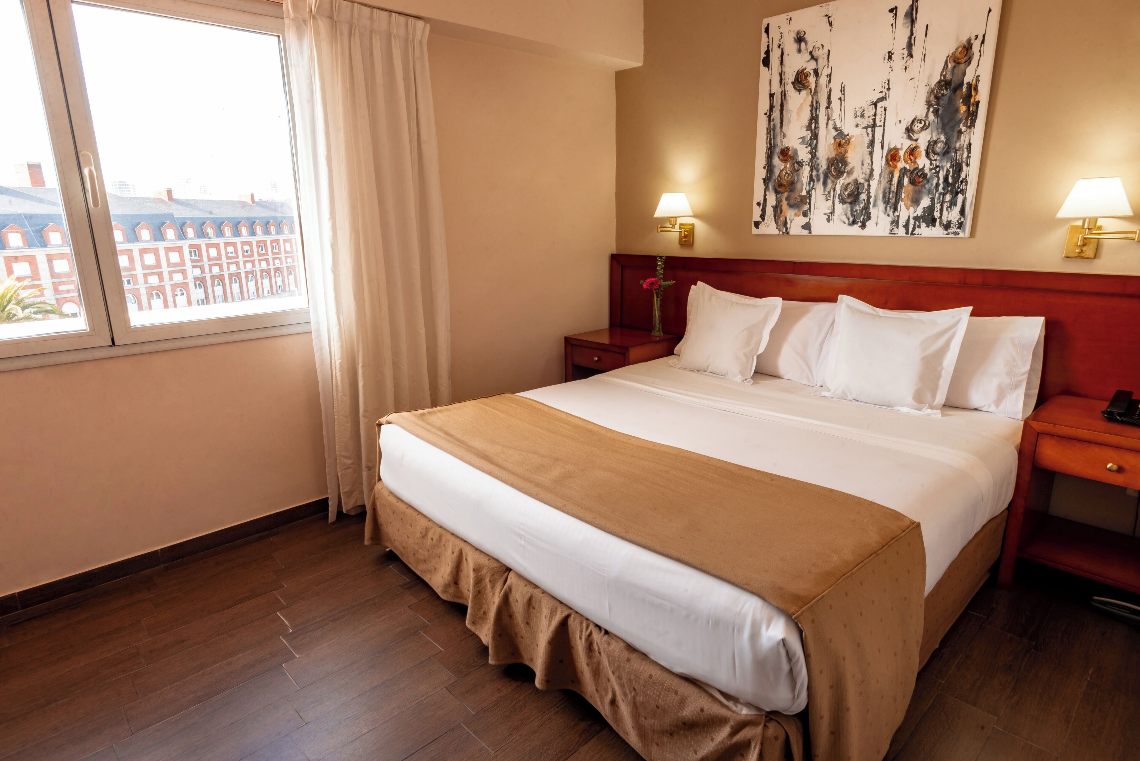 A hotel room with a large bed, a nightstand with a lamp, a painting on the wall, and a window with curtains.