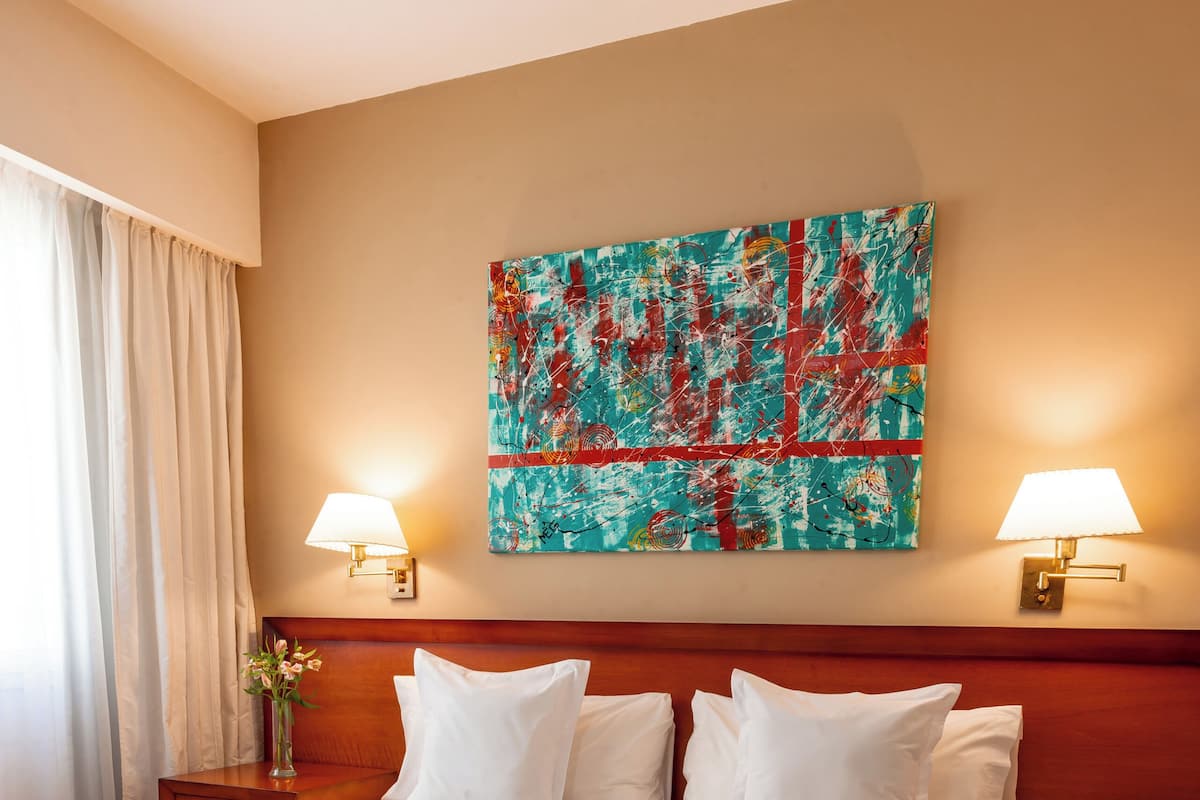 Une chambre d’hôtel avec un grand lit, deux tables de chevet, une petite table, un tableau accroché au mur et une fenêtre avec des rideaux.