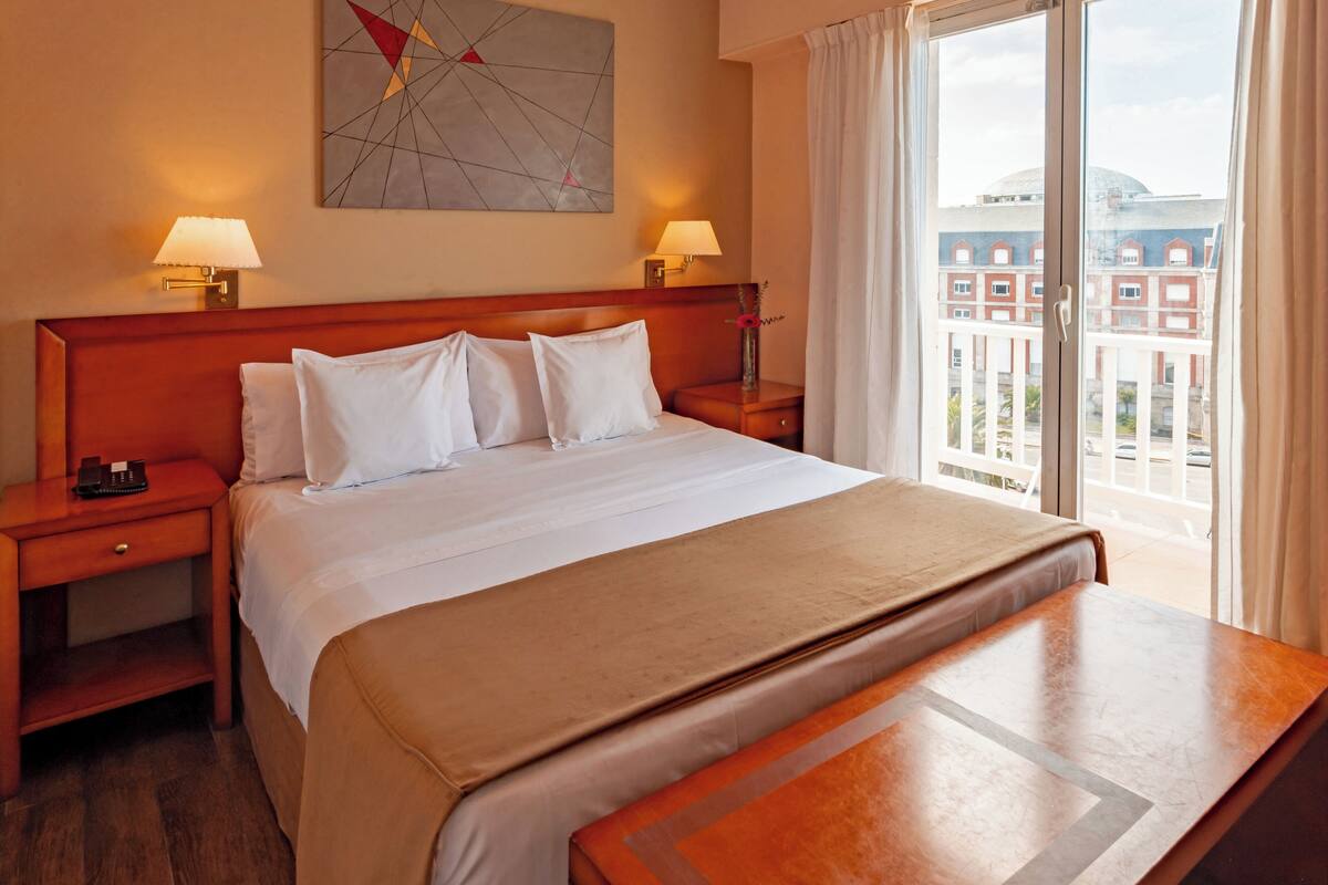 Une chambre d’hôtel avec un grand lit, deux tables de chevet, une petite table, un banc en bois, un tableau et une vue sur les bâtiments par la fenêtre.