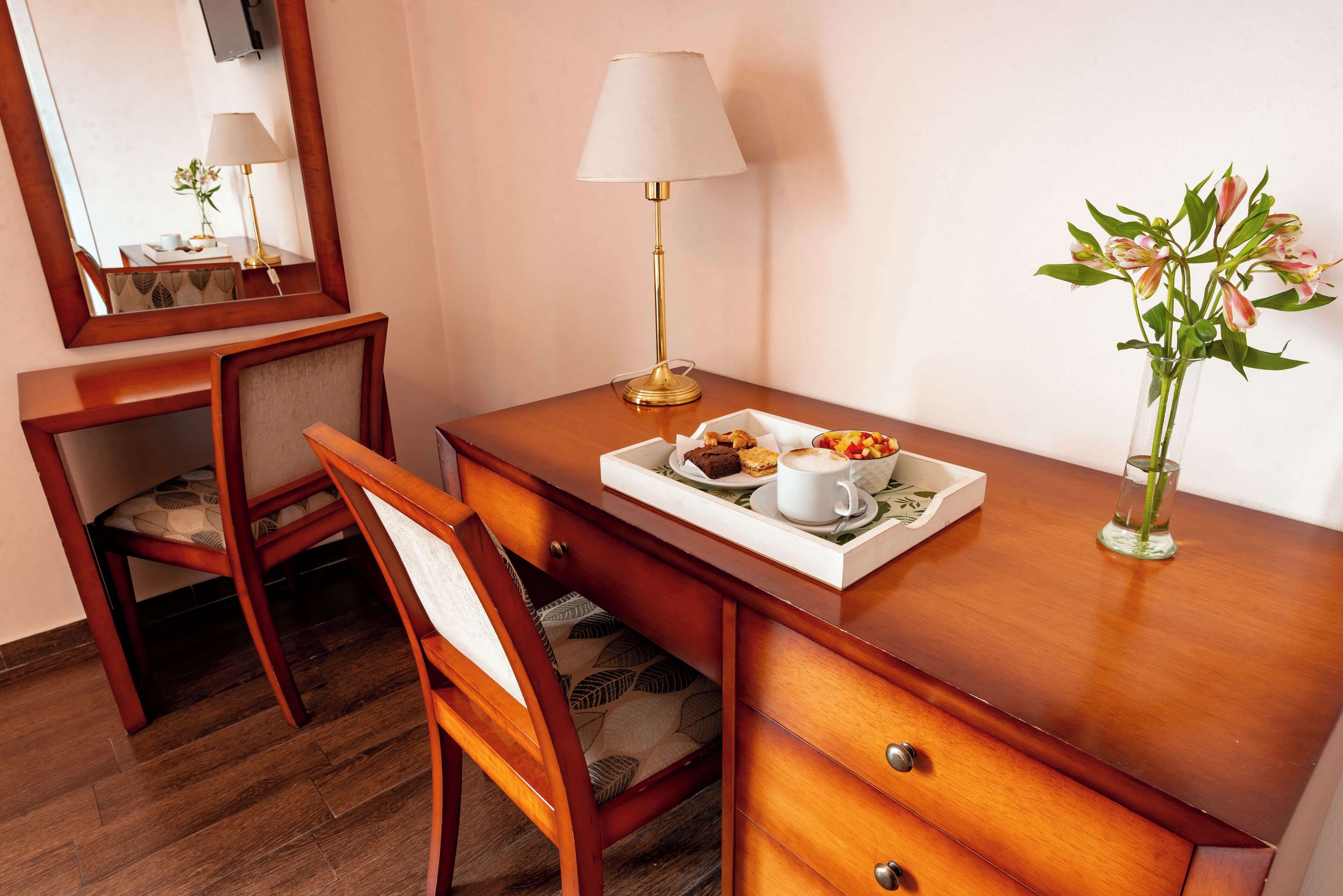 A wooden desk with a lamp, a vase with flowers, a tray with food and a cup, and a chair.