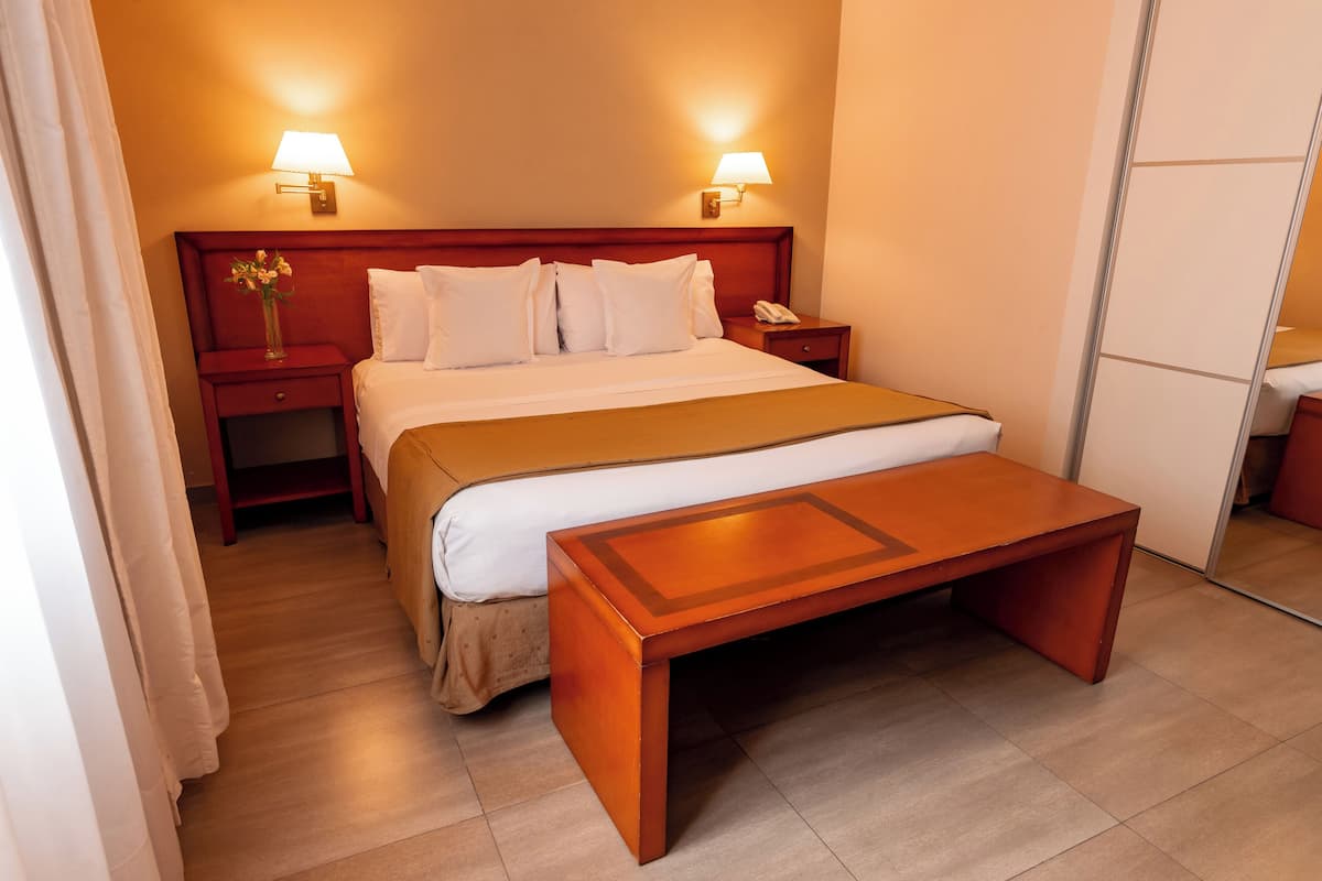 Une chambre d’hôtel comprenant un lit, des tables de chevet, une petite table, un banc en bois et deux lampes fixées au mur.