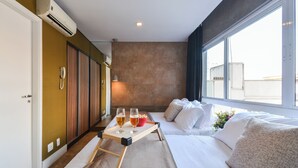 A hotel room with a bed, a bedside table with glasses and a bowl of cereal, a window with a view of buildings, and an air conditioning unit. - Studio with Pool and Garage – 7 min from Allianz (São Paulo)
