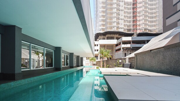 A modern indoor swimming pool with clear blue water, surrounded by a minimalist design with large windows and a view of a high-rise building under construction. - Studio with Pool and Garage – 7 min from Allianz (São Paulo)