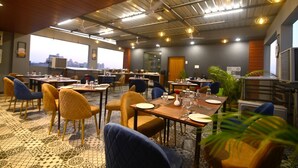 Un comedor de hotel con varias mesas y sillas, un techo alto con vigas a la vista y grandes ventanales que ofrecen vistas a la ciudad.