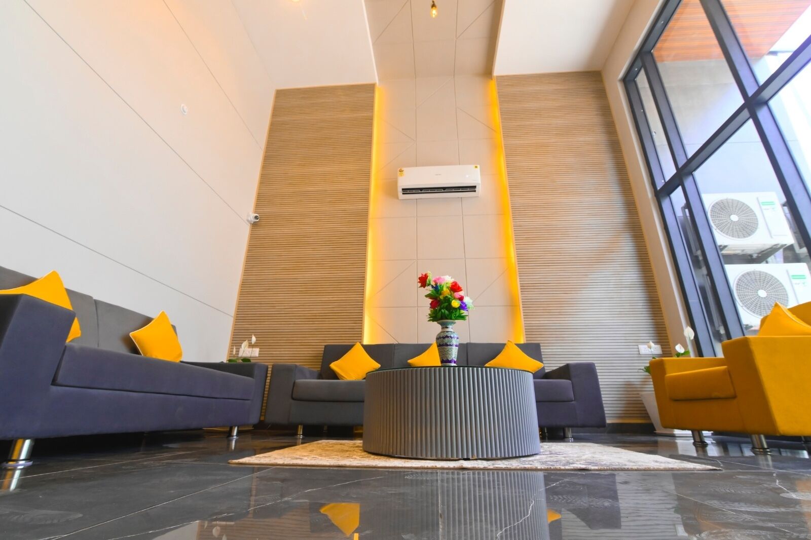 Een moderne lobby met een grijze bank, een ronde salontafel met een bloevas en een groot raam met airconditioningsapparaten.