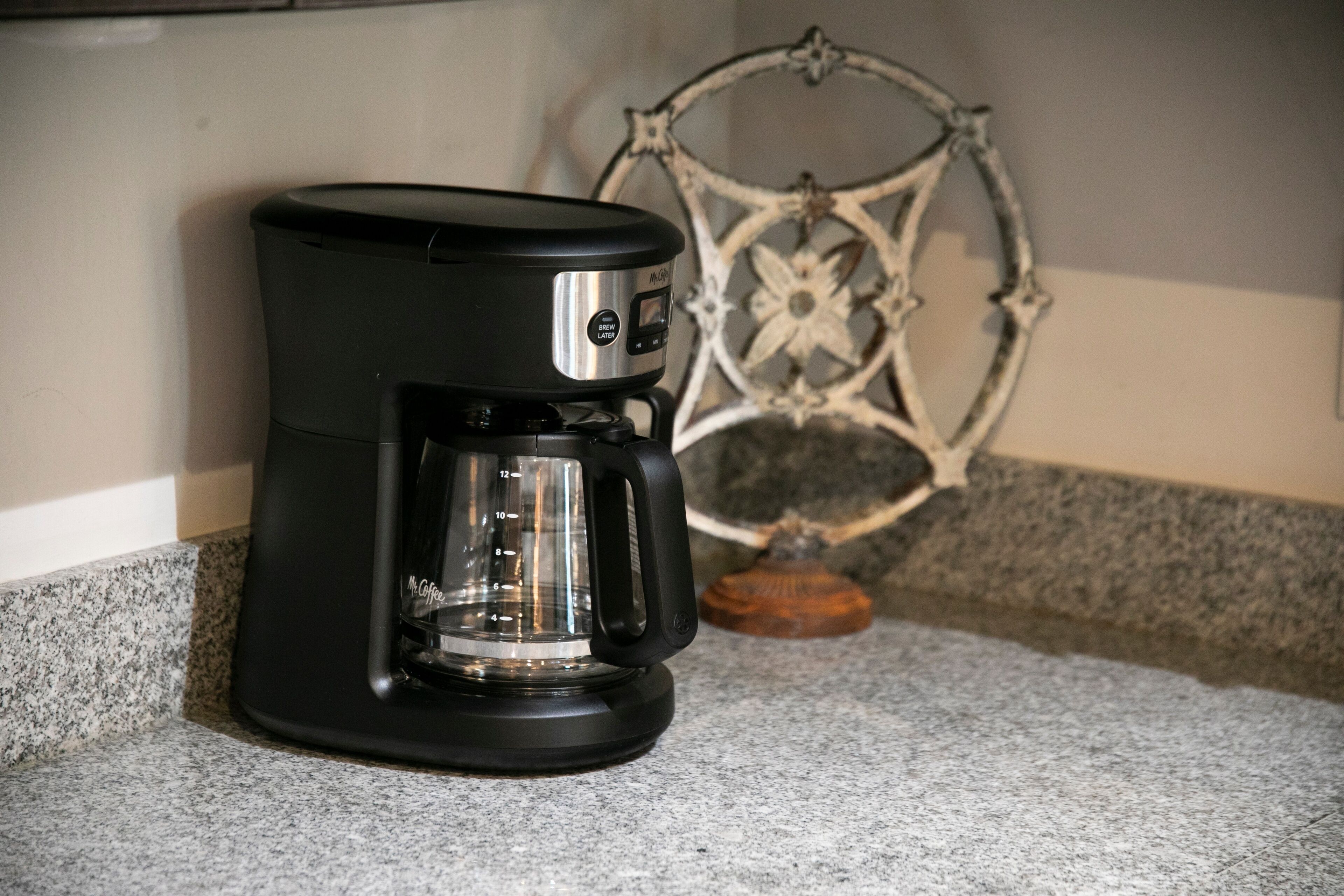 Une machine à café noire posée sur un comptoir en granit, avec un élément décoratif en métal en arrière-plan.