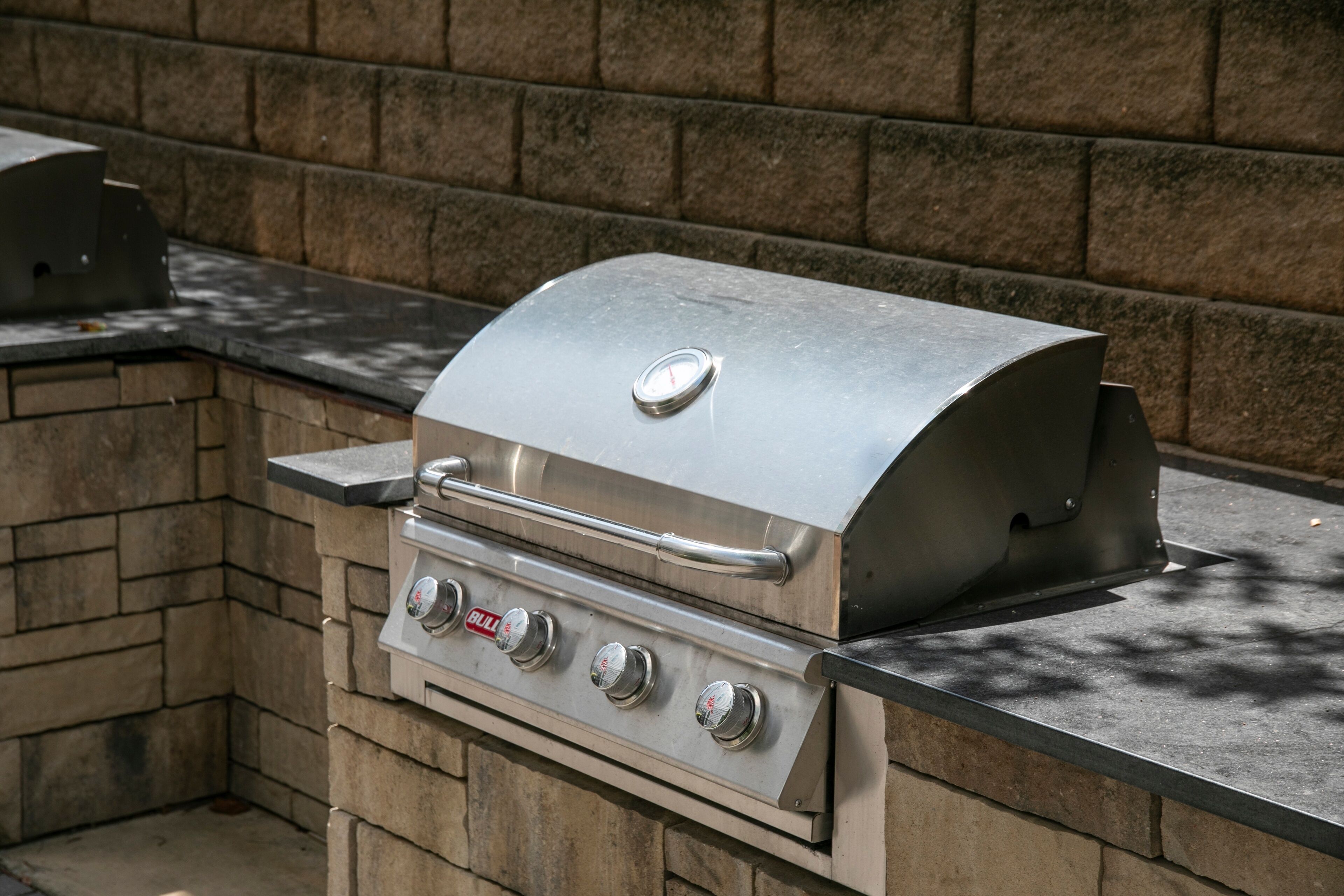 A stainless steel grill with four knobs and a lid, set against a stone wall and a dark countertop.