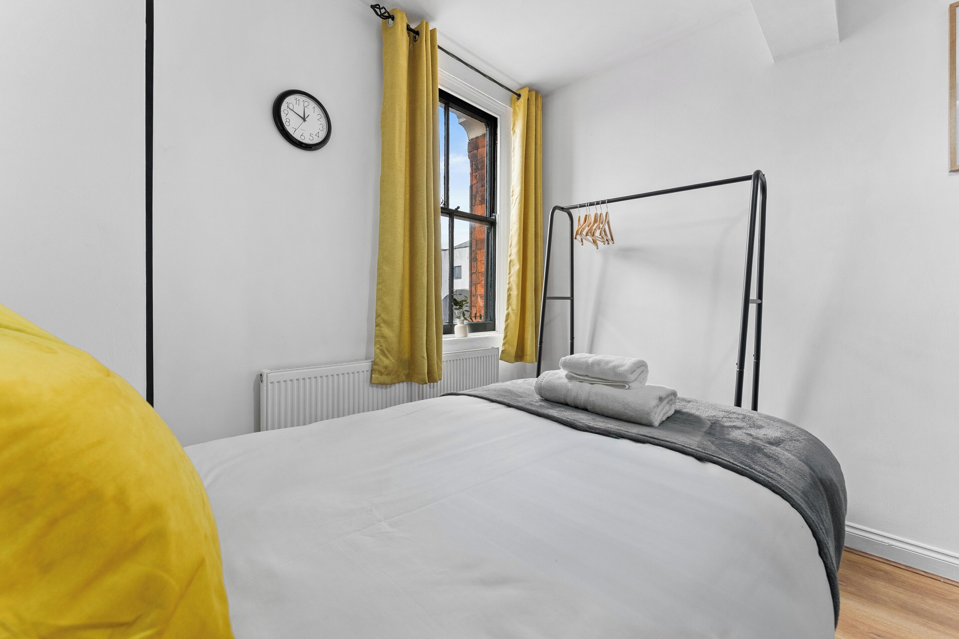 A modern bedroom with a bed, a window with yellow curtains, a radiator, and a clock on the wall.